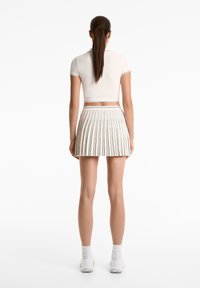 Bílý zkrácený polo s krátkými rukávy, doplněný o bílou plisovanou sukni s vertikálními pruhy. Modelka stojí zády k nám a má na sobě tenisky.