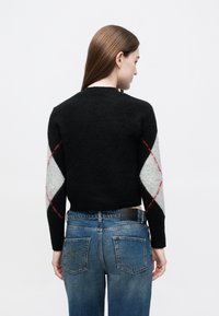 Črn pulover z teksturirano površino, z rokavi v sivi in beli argyle vzorcu, poudarjenimi z rdečimi črtami. Nošen z modrimi kavbojkami iz jeansa.