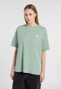 T-shirt en coton vert menthe à manches courtes, coupe décontractée, col rond, avec un détail de logo blanc sur la poitrine. Jean noir visible en dessous.