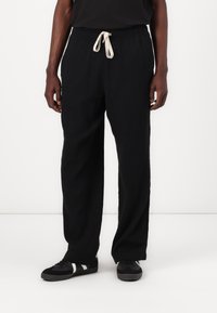 Pantalons noirs amples avec une taille élastique et un cordon beige, en tissu texturé. Portés avec des chaussures noires avec des accents blancs.