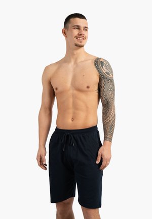 Hemdloser Mann mit kurzen Haaren und einem Tribal-Tattoo mit vollem Ärmel am linken Arm trägt schwarze Shorts, lächelt und schaut zur Seite.