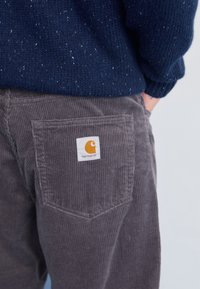 Szare spodnie kordowe z tylną kieszenią, zdobioną białą łatą z logo. Tkanina ma żebrową fakturę, dopełnioną ciemnoniebieskim swetrem dżersejowym.