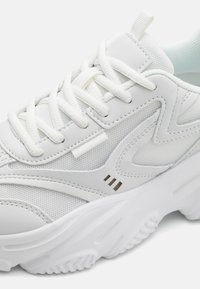 White athletic shoe with a mix of mesh and synthetic leather materials. Features white laces, textured sole, and decorative side accents.