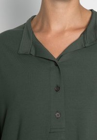 Polo de manga corta verde oscuro con cuello, tres botones negros y una textura suave. Incluye detalles de costura sutiles.