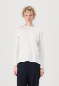 Langärmliges weißes Baumwoll-T-Shirt mit Rundhalsausschnitt und leicht abgerundetem Saum. Weicher Stoff, lockere Passform, über dunkler Hose getragen.