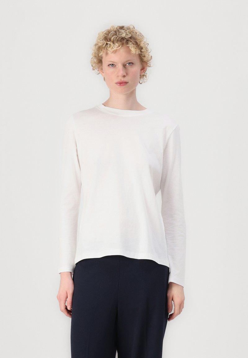 Langärmliges weißes Baumwoll-T-Shirt mit Rundhalsausschnitt und leicht abgerundetem Saum. Weicher Stoff, lockere Passform, über dunkler Hose getragen.
