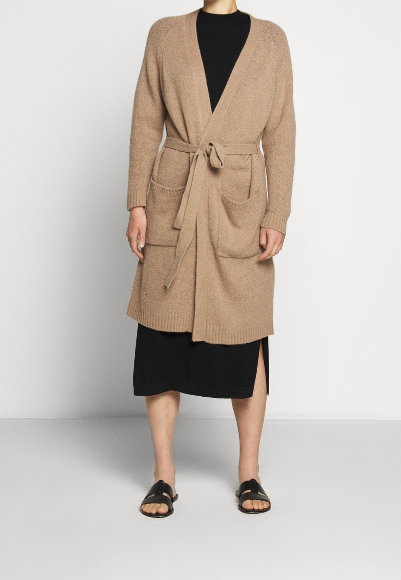 Kobieta w beżowym kardiganie z paskiem i dużymi kieszeniami, nosząca czarną sukienkę midi oraz czarne sandały na płaskiej podeszwie, stojąca na tle w jednolitym kolorze.