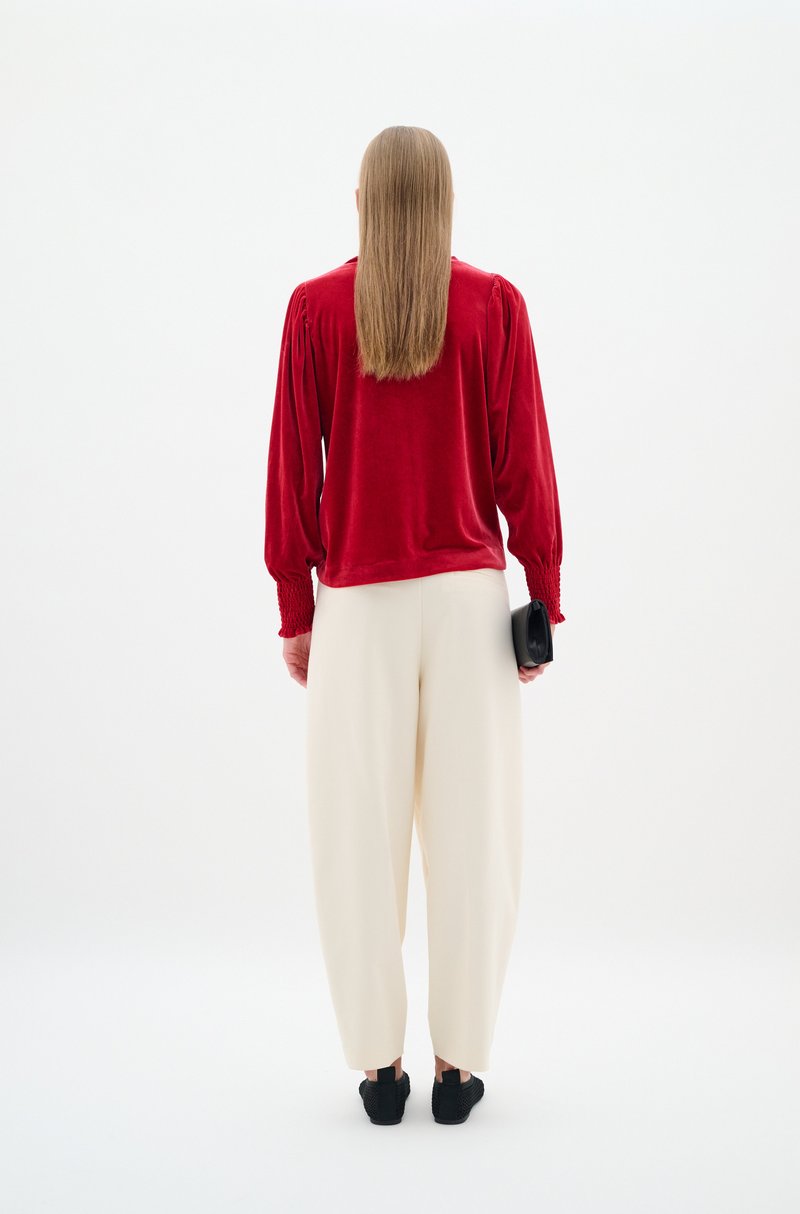 Langarmoberteil aus rotem Samt mit gerafften Manschetten, kombiniert mit kremfarbenen, weit geschnittenen Hosen. Eine schwarze Clutch und strukturierte Schuhe runden das Outfit ab.