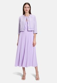 Krótkie lilakowe żakiet z tkaniny tweedowej, z rękawami do łokcia, ozdobiony złotymi guzikami, noszony na zwiewnej, lilakowej sukience plisowanej. Prosta, miękka tekstura.