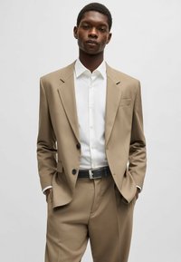Beige pak met een gestructureerde blazer, wit overhemd met knopen en een zwarte riem. Opvallende kenmerken zijn twee knopen en een voorzak.