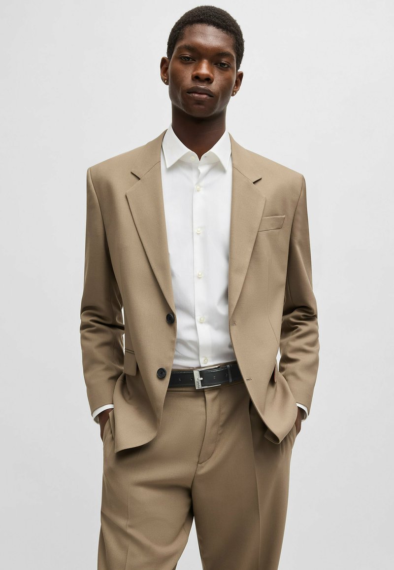 Beige pak met een gestructureerde blazer, wit overhemd met knopen en een zwarte riem. Opvallende kenmerken zijn twee knopen en een voorzak.