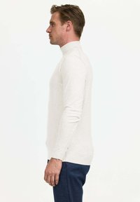 Pull col roulé gris clair avec de longues manches, coupe ajustée, fabriqué dans un matériau doux, associé à un pantalon texturé bleu foncé.