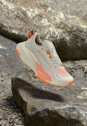 Bēšs un oranžs The North Face taku skriešanas apavu pāris, kas it kā lido virs klinšainas virsmas, paredzēts lietošanai brīvā dabā un nelabvēlīgos apstākļos.