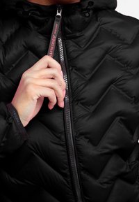 Giacca a vento nera con motivo a trapunta a zigzag e colletto alto. Presenta una cerniera metallica con un tira-zip con marchio.
