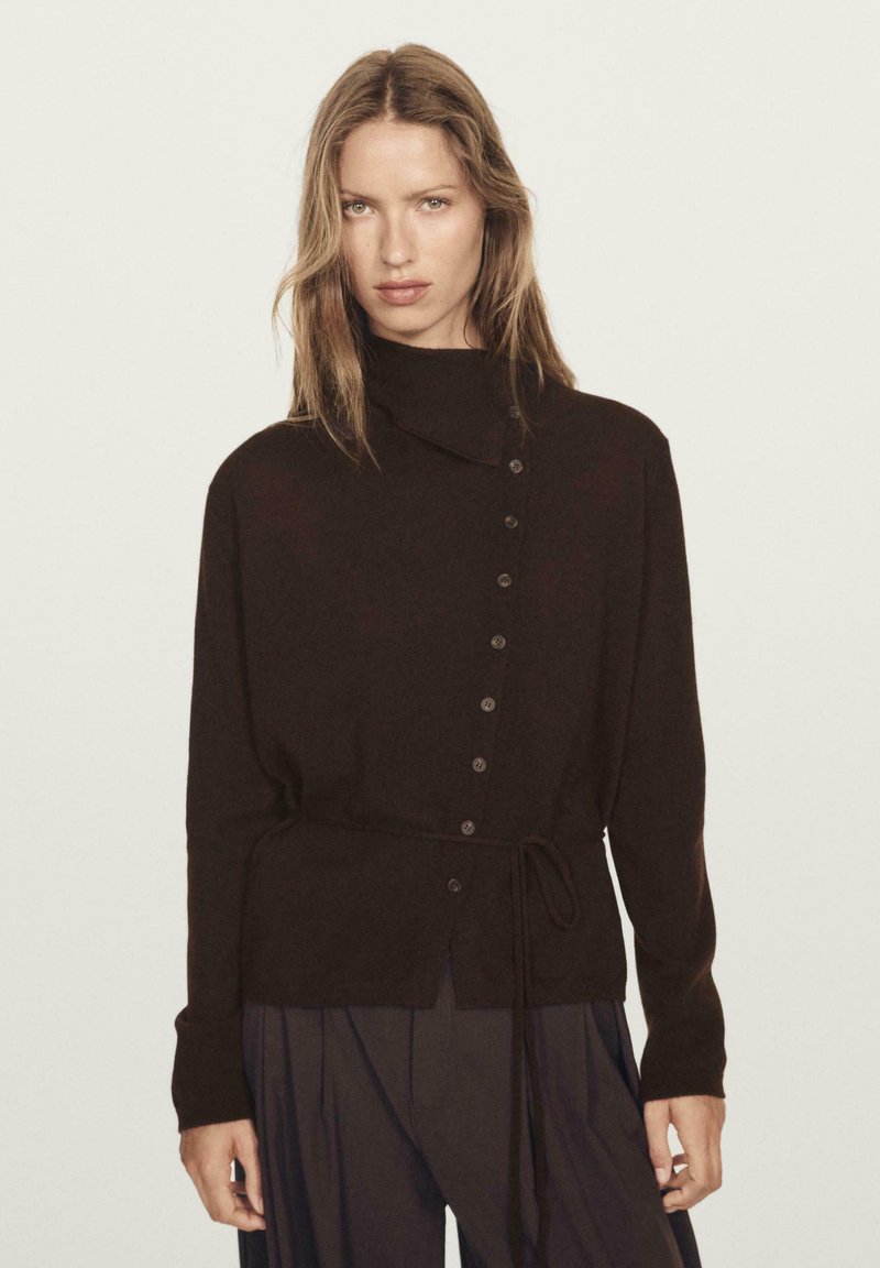 Maglione a maglia marrone scuro con colletto alto, bottoni frontali e dettagli con vita legata. Le maniche lunghe e la vestibilità morbida completano il design.