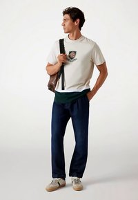 Férfiaknak szánt hétköznapi öltözék, amely krémszínű pólót tartalmaz zöld logóval, sötét farmernadrágot és bézs színű, zöld részletekkel díszített sportcipőt, barna válltáskát tartva.