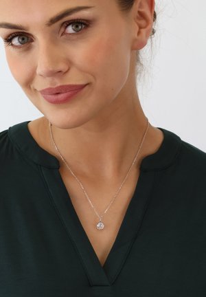 Eine silberne Halskette mit einem detaillierten Anhänger hängt an einer zarten Kette. Die Trägerin trägt ein dunkelgrünes Oberteil mit V-Ausschnitt und hat ein natürliches Make-up.