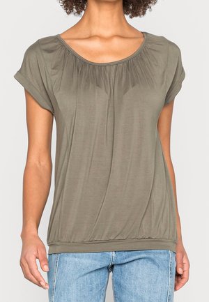 Frau trägt ein locker sitzendes, kurzärmeliges olivgrünes Oberteil mit gerafftem Ausschnitt und Saum, kombiniert mit hellblauen Jeans.