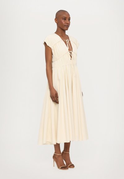 Lichtbeige midi-jurk met korte capmouwen, trekkoordhalslijn en geplooid middel. Gecombineerd met nude hoge hakken.
