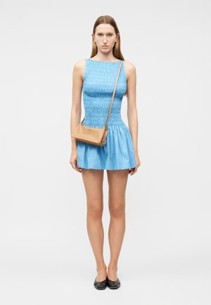 Γυναίκα που στέκεται και βλέπει μπροστά, φορώντας ένα αμάνικο μπλε φόρεμα, μπεζ τσάντα χιαστί και μαύρα επίπεδα παπούτσια σε ένα απλό λευκό φόντο.