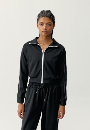 Žena s vrkočmi, ktorá má na sebe čiernu bundu so zipsom a čierne nohavice so sťahovacou šnúrkou a bielym lemovaním na jednofarebnom pozadí.