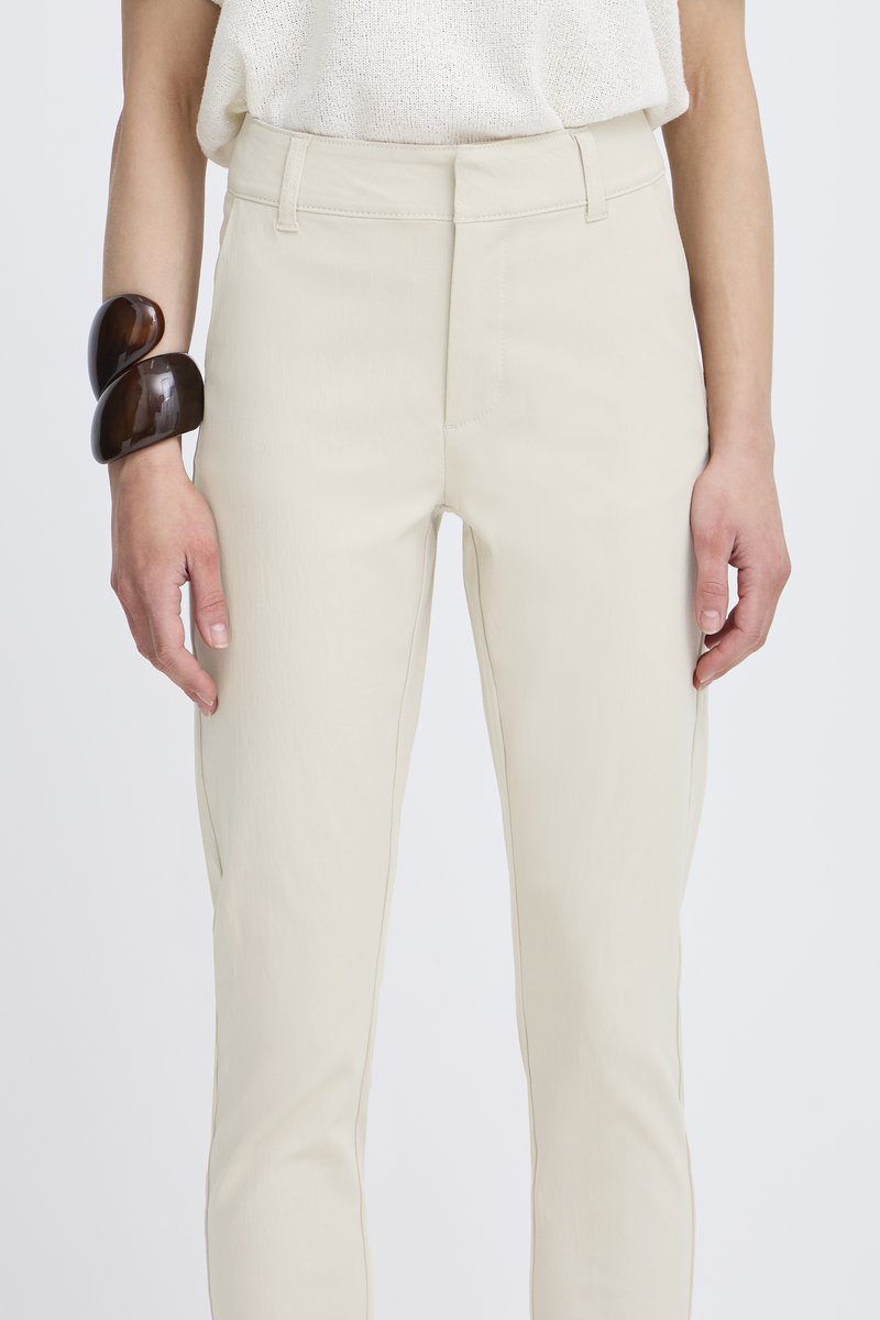 Beige slim-fit kalhoty vyrobené z lehkého materiálu s hladkou strukturou. Na zápěstí je ozdobný náramek tmavohnědé barvy.