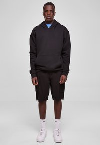Felpa nera con cappuccio e tasca a canguro, abbinata a pantaloni cargo neri fino al ginocchio. Sneakers bianche con dettagli neri e calzini alti.