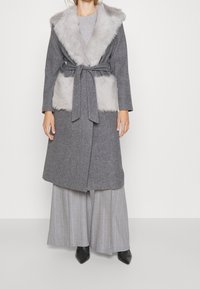 Woman wearing belted gray wool coat with light gray faux fur collar and pockets, paired with wide-leg striped gray pants and black pointed boots.