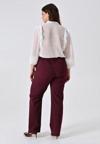 Donna in una camicia bianca trasparente con maniche a volant e pantaloni bordeaux a vita alta, in piedi contro uno sfondo semplice.