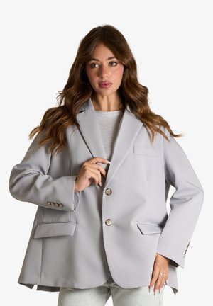 Donna con lunghi capelli ondulati castani che indossa un blazer oversize grigio chiaro sopra una maglia bianca e jeans azzurri chiari su sfondo bianco.