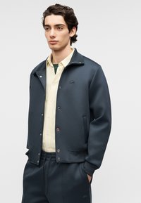 Chaqueta azul marino con botones de presión, con un logo sutil. Llevada sobre una camisa de cuadros amarillo claro, combinada con pantalones deportivos a juego.