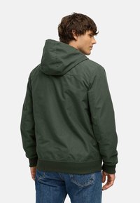 Groene hooded jas met een gladde, matte afwerking, voorzien van een geribde zoom en manchetten. Gekozen met blauwe jeans, wat een ontspannen pasvorm laat zien.