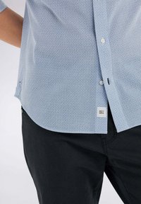 Camicetta azzurro chiaro a maniche corte con un piccolo motivo geometrico bianco e blu, presenta un colletto, bottoni e un'etichetta che recita "KEEP IT SIMPLE."