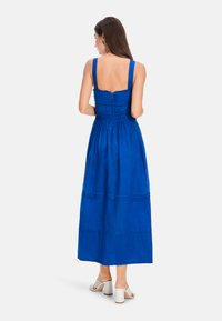 Blaues Maxikleid mit breiten Trägern, gerafftem Oberteil und gestuftem Design. Reißverschluss auf der Rückseite und fließender Stoff, kombiniert mit weißen Blockabsatzsandalen.