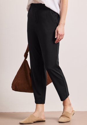 Kvinde iført sorte skrånende bukser, beige slip-on sko og holder en brun ruskindtaske mod en neutral baggrund.