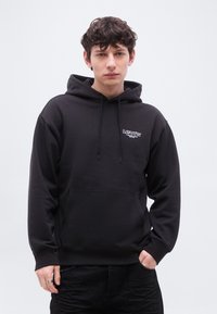 Νέος άνδρας με σγουρά μαλλιά, φορώντας μαύρο φούτερ Levi’s με μπροστινή τσέπη και λευκό λογότυπο στο στήθος, στέκεται μπροστά σε απλό φόντο.