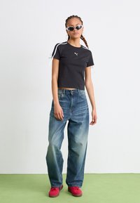 Tricou scurt negru cu accente albe pe laterale, asociat cu blugi largi albaștri. Modelul poartă pantofi roșii și ochelari de soare albi. Fundal simplu.
