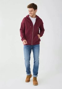 Muškarac u bordo zip-up hoodie, bijelom majici, plavim trapericama i smeškim čizmama boje tamne bež, stoji i smeška se protiv bijele pozadine.