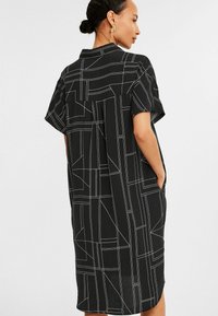 Schwarzes Kurzarmkleid mit geometrischem weißen Linienmuster, lockerer Passform und Seitentaschen. Leichter Stoff mit detailierter Kragen.