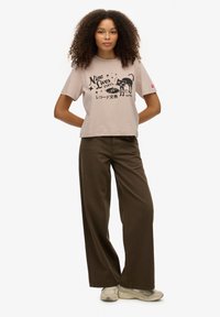 Lyserød bomulds-T-shirt med sort grafisk print, korte ærmer og løs pasform. Sammen med mørkebrune bukser med brede ben og beige sneakers.