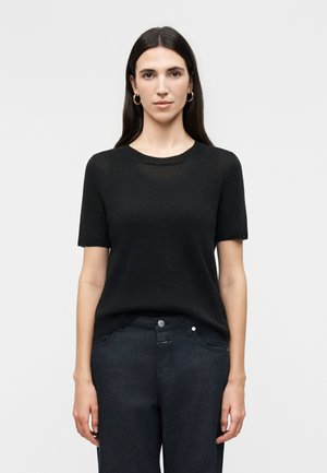 Γυναίκα με μακριά σκουρόχρωμα μαλλιά που φορά χρυσούς κρίκους στα αυτιά, μαύρο πουλόβερ με κοντά μανίκια και σκούρο μπλε τζιν, στέκεται μπροστά σε ένα απλό φωτεινό φόντο.