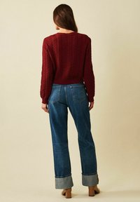 Burgundyröd texturerad stickad tröja med långa ärmar, i kombination med högmidjade blå denimjeans med uppvikta benslut och beige klackskor.