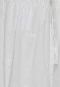 Vêtement en coton blanc avec une broderie florale subtile, taille élastique froncée et fermeture à lien sur le côté. Texture douce et légère.