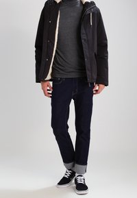 Svart jacka med fårskinnsfoder, mörkgrå polotröja, mörkblå jeans och svarta sneakers med vita sulor, med upprullade ärmar.