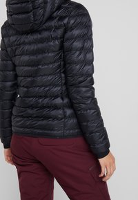 Svart puffjacka med quiltad design, huva och elastiska ärmar. Bärs över vinröda byxor som visar en slät, glänsande textur.