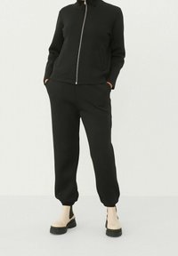 Survêtement noir zippé avec col montant ; coupe ajustée, manches longues et pantalon style jogger ; texture lisse ; porté avec des bottines noires et beiges.