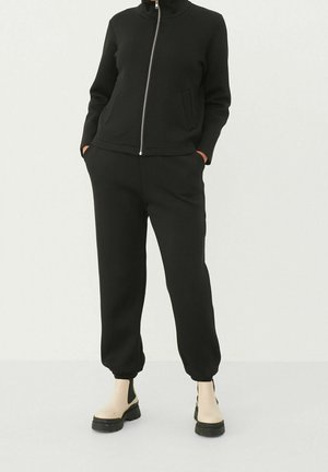 Svart zip-up-träningsdräkt med hög krage; åtsittande design, långa ärmar och joggerbyxor; slät textur; bärs med svarta och beigea ankelstövlar.