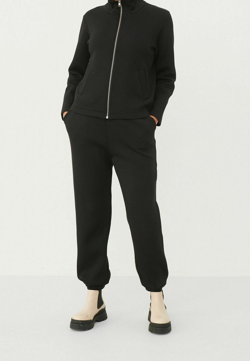 Survêtement noir zippé avec col montant ; coupe ajustée, manches longues et pantalon style jogger ; texture lisse ; porté avec des bottines noires et beiges.