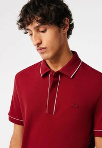 Röd pikétröja med en subtil lyster, med en tvåknappsslå, vit piping längs kragen och ärmarna, samt en liten logotyp på bröstet.