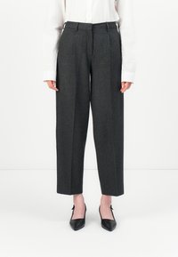 Personne portant un pantalon tailleur gris foncé cropped, une chemise blanche à manches longues et des escarpins noirs à bout pointu, se tenant sur un fond blanc.